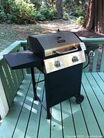 Front left side view of the Dyna-Glo barbecue grill on a green porch showing wheels and side shelf.