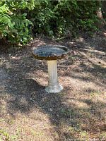 Concrete bird bath standing outdoors on soil with visible wear and chips on base and rim; black metal bird feeder cage sitting in top basin.