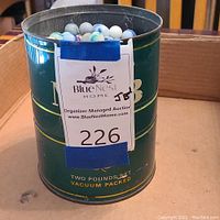 Photo showing the green metal coffee can with blue tape and auction label on it, marbles inside visible at top edge.