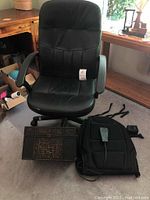 Black leather desk chair with armrests and caster wheels, Homedics portable chair massager, and rattan wood decor box placed on floor in natural light.