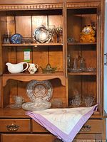 Wooden hutch with shelves displaying various glassware including plates, juice squeezer, creamers, mini milk bottles, and an embroidered tablecloth on the drawer surface