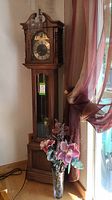 Full view of tall wooden grandfather clock with glass front and moon phase dial, standing next to window with flowing curtain and vase of faux flowers in front