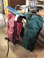 Red Jansport hiking backpack with visible wear and a green camp chair in carrying bag beside it, placed on concrete floor.