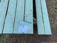 Top view of the green-painted wooden picnic table surface showing six wide planks with visible scratches and two benches on either side, one partially missing. A MaxSold tag in a plastic bag rests on the table.