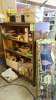 Wooden shelf with multiple shelves filled with painting and home improvement supplies and a boxed Sears tent leaning against it.