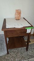 Wooden side table shown with white cloth on top and a rock crystal lamp standing on it. The table has a lower shelf and visible scratches.