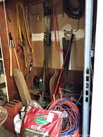 Wide view showing tools hanging on pegboard and wall including large tree saw, hand saws, bow saw, sledgehammer, extension cords, red gas can, and various other tools and supplies.