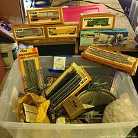 Multiple boxed Tyco model train cars displayed on wood surface above plastic bin with track pieces and wiring parts inside.