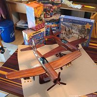 Two wooden model airplanes displayed on a table with five boxed jigsaw puzzles behind them. The larger plane is in front, showing polished wood, detailed cockpit, and rotating propeller. Puzzles visible include 'Seaport Marketplace' and floral 500-piece.