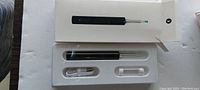 Black wireless ear cleaning pod in packaging with USB charger and two spare head attachments visible.