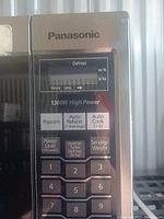 Close-up of control panel showing Panasonic brand, cooking options such as Popcorn, Auto Reheat, Auto Cook, Defrost with settings for weight and power level, numeric keypad and start/stop buttons.