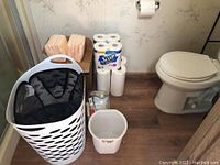 View showing two white laundry baskets, small white wastepaper bin, four packages of beige medical pads, and 20 rolls of Scott toilet paper stacked near a toilet.