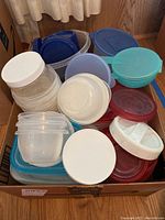 Photo showing assorted plastic containers and lids in a cardboard box, various sizes and colors including white, blue, and red.