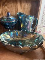 Three blue-green carnival glass pieces: pitcher, lidded candy dish, and bowl displayed on wood shelf.