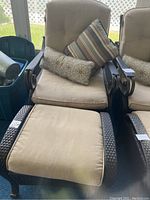 Overall view of chair and matching stool with cushions showing brown plastic rattan weave and beige cushions with patterned pillows.
