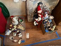 Wide shot of over 20 Christmas decorations and the large pillow, ladder decoration, many snowmen and Santa figures scattered on plastic surface and in box, capturing the lot contents.