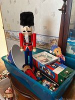 Large wooden nutcracker with black fur hat standing beside a blue plastic container filled with smaller nutcrackers and a boxed item, showing scale and condition.