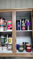 Cabinet shelf with assorted coffee mugs and travel mugs including Coca-Cola and Chicago Cubs themed cups, Grandpa and Grandma mugs.