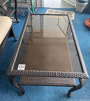 Top view of rectangular glass top coffee table with rattan-wrapped frame and lower woven plastic rattan shelf, placed on blue carpet.