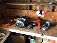 Photo of Remington electric chain saw, Fury drill, Craftsman hand saw and belt sander on wooden shelf with orange extension cord.