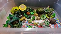 Overview of assorted ceramic and plastic frog figurines in a plastic bin, various sizes and colors, need cleaning.