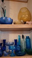 Top and middle shelf with amber and blue vases, bowls, and decorative glass. Includes a large smoky amber ribbed vase, amber scalloped bowl, and various blue glass bottles and vases.