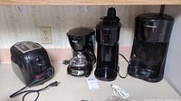Four black small kitchen appliances including toaster, drip coffee maker, coffee grinder, and coffee maker on kitchen counter