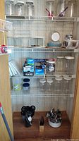 Wide view of kitchen storage shelf with various clear plastic containers with white lids, kitchen plates, boxed plastic wrap and storage bags, flatware, and utensils.
