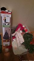 Photo showing boxed 32-inch Christmas tree alongside various decor including ribbons, Santa caps, tree skirt, and base of tree.