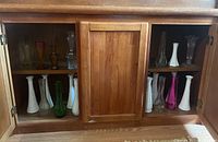 Multiple bud vases made of white milk glass, clear glass, and green glass arranged inside a wooden cabinet.