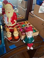 Vintage light-up Santa figurine standing on a dresser next to two choir angel candle holders and a box of assorted bulbs, as well as the ceramic elf sitting at the edge of the dresser.