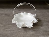 Front view of white Fenton Silver Crest bridal basket with clear glass ruffled edge and handle on wooden surface.