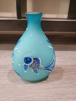 Front and side view of teal blue satin glass vase with a detailed hand painted blue and gold fish, showing the texture and design on the vase body.