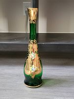Front view of emerald green glass vase with gold and enamel floral decorations on a dark shelf.