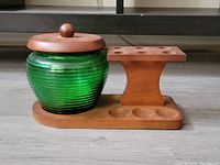 Side view of green ribbed glass humidor jar with fitted wooden lid and adjacent wooden pipe rack on same base.