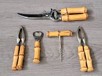 Four bar tools arranged on a flat surface: curved serrated knife, nutcracker, bottle opener, winged corkscrew. All handles made of bamboo-style wood with chrome metal parts.
