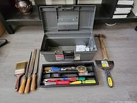 Rubbermaid gray plastic tool box opened with removable tool tray partially filled with assorted screwdrivers, pliers, tape measure, and other small hand tools; paintbrush, three files with wooden handles, hammer with wooden handle, and yellow and black putty knife placed next to box