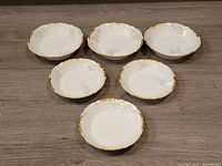 Top view of 6 white porcelain bowls with hand painted pastel flowers and gold gilding around scalloped rims, arranged on wooden surface.