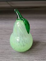 Front view of the speckled light green art glass pear with green stem and leaf, placed on a wooden surface.