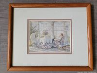 Framed print with visible wooden frame, white matting, and original pink back mat showing. The print depicts a woman sitting in a floral armchair near a table with baby carriage and basket.
