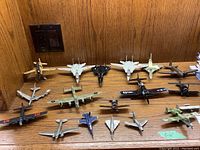 Seventeen cast metal miniature planes arranged on wooden surface against wood backdrop. Various colors and styles including WWII era propeller planes and modern jet fighters. Visible wear and chipped paint on several pieces.