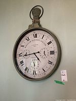 Front view of the large oval wall clock hanging on a plain wall, showing the clock face with roman numerals and three sub-dials, a rope loop handle on top, and the brand and date markings.