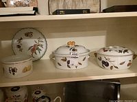 Photo showing four Evesham ceramic bakeware items on shelf, including a dish with a lid and sculpted fruit handle, a round ribbed dish with fruit design, and additional casserole dishes.