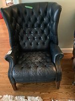 Front view of black leather wing back chair showing tufted back and heavily worn seat with visible cracking, nail head trim on arms.