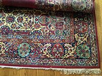 Rolled rug showing red guard border, floral motifs, and cotton fringe at one end