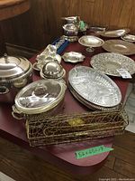 Wide view of silver plated serving ware including dishes, gravy boat, water jug, butter dish, centerpiece, floral frogs, and salt and pepper shakers arranged on a table.