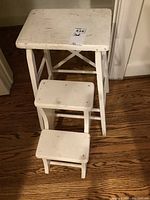Front angled view of three vintage white painted wooden step stools of different sizes on hardwood floor.