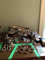 Wide view of the four sterling silver napkin rings, two tiny sterling salt spoons, and additional silver plate accessories arranged on a wooden surface.