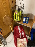 Full lot showing wooden racquet, Wilson ball hopper filled with yellow balls, Wilson red racquet bag, and used balls.