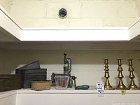Shelved lot items including 4 brass candlesticks, metal meat grinder, Pyrex measuring cup, metal loaf pans, round speckled pan, French fry cutter.